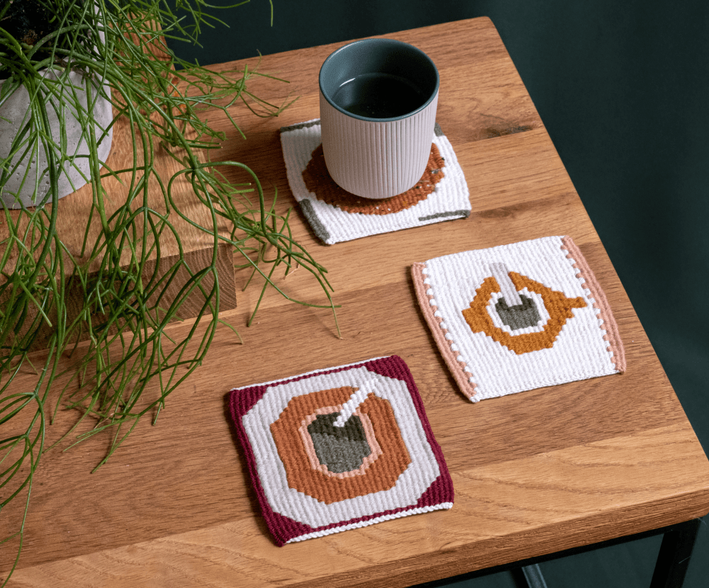Three handwoven coasters with a mate-cup motif, on a wooden table beside a cup of tea